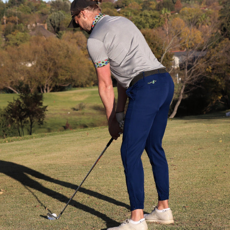 Man playing golf on a grassy course with trees and bushes in the background. Wearing golf joggers.