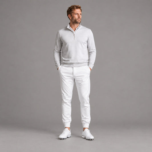 A man modeling a light gray quarter-zip pullover sweater and white jogger pants with white golf shoes, standing against a solid gray studio background.