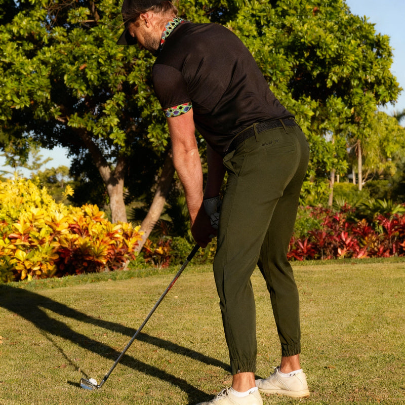 Man playing golf on a grassy course with trees and bushes in the background. Wearing golf joggers.