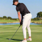 Man playing golf on a course with trees in the background, wearing jogger style trousers in white.
