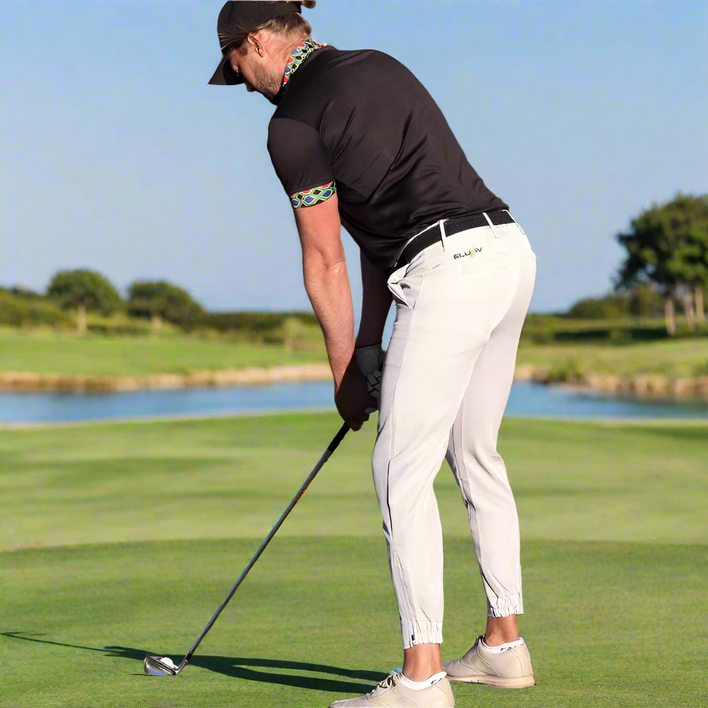 Man playing golf on a course with trees in the background, wearing jogger style trousers in white.