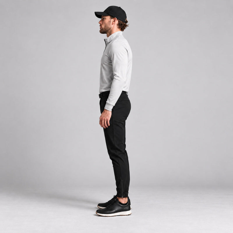 A man in profile wearing a light gray long-sleeve quarter-zip pullover, black slim-fit pants, black sneakers, and a black baseball cap, standing against a neutral light gray studio background.