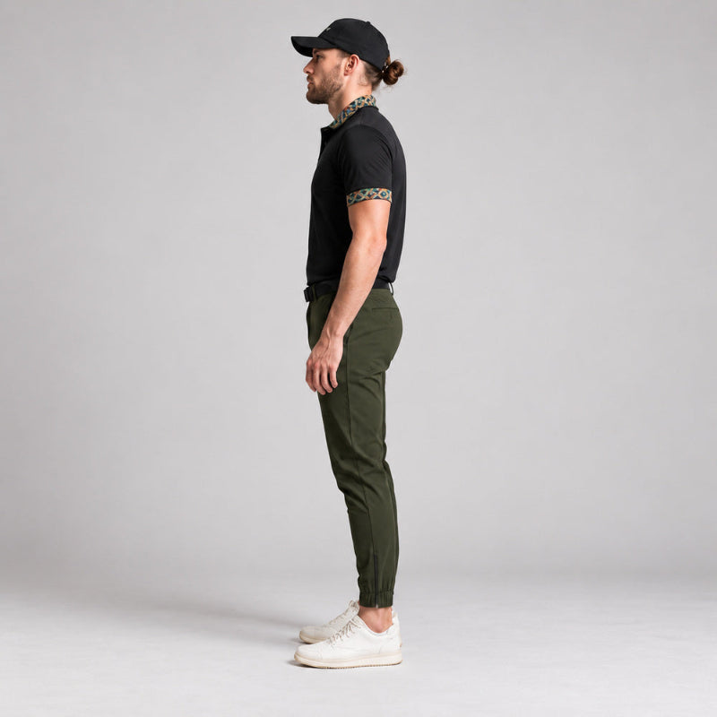 A man in profile wearing a black short-sleeve polo shirt with patterned trim, olive green slim-fit trousers, and white sneakers, standing against a light gray studio background.