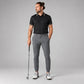 A man modeling a black short-sleeve polo shirt with patterned trim on the collar and sleeves, paired with grey trousers and white golf shoes, holding a golf club against a light grey studio background.