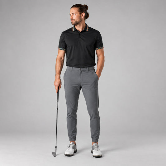 A man modeling a black short-sleeve polo shirt with patterned trim on the collar and sleeves, paired with grey trousers and white golf shoes, holding a golf club against a light grey studio background.
