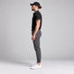 A man in profile wearing a black short-sleeve polo shirt with patterned trim on the collar and sleeve, dark grey slim-fit trousers, and white sneakers, standing against a light grey studio background.