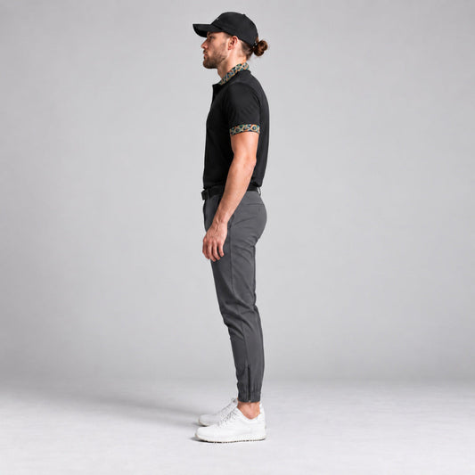 A man in profile wearing a black short-sleeve polo shirt with patterned trim on the collar and sleeve, dark grey slim-fit trousers, and white sneakers, standing against a light grey studio background.