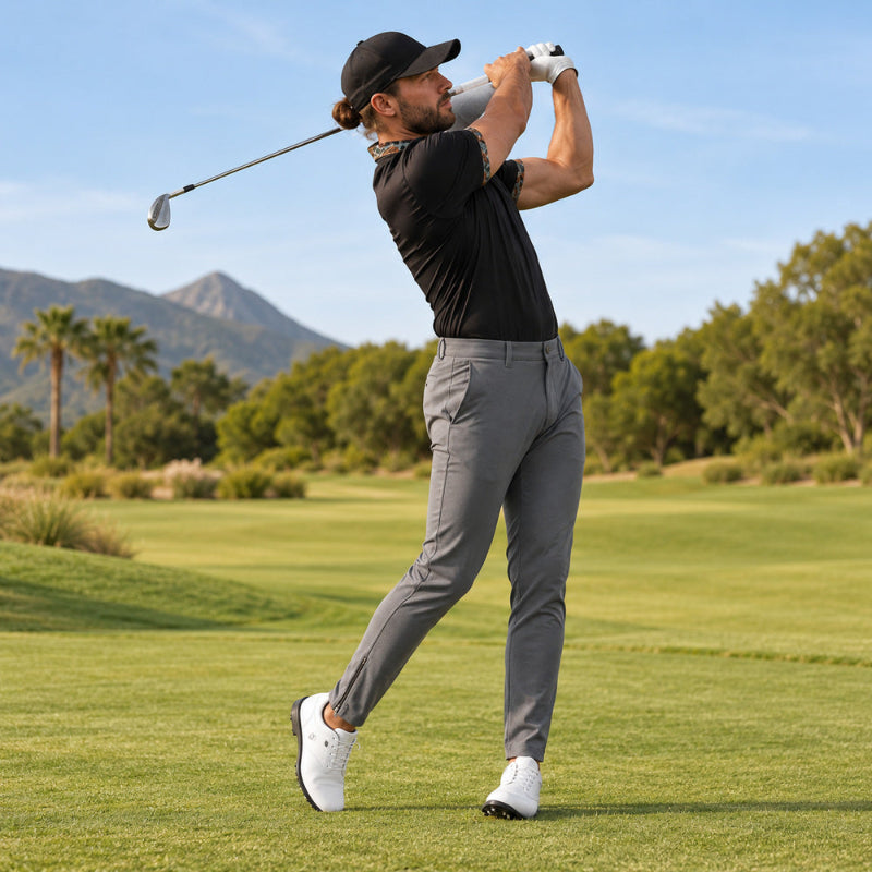 A man in a black polo shirt, grey trousers, white golf shoes, and a black cap is captured mid-swing on a golf course with green grass, trees, and mountains in the background.
