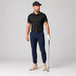 A male model stands on a light gray studio background wearing a black short-sleeve polo shirt with patterned trim on the collar and sleeve cuffs, navy golf pants, a white golf glove, beige golf shoes, and a black cap. He is holding a golf club.