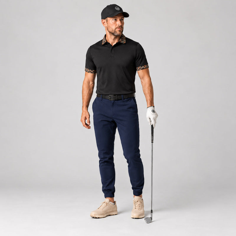 A male model stands on a light gray studio background wearing a black short-sleeve polo shirt with patterned trim on the collar and sleeve cuffs, navy golf pants, a white golf glove, beige golf shoes, and a black cap. He is holding a golf club.