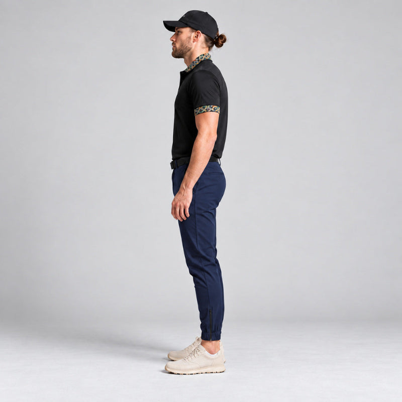 A man in profile wearing a black short-sleeve polo shirt with patterned trim on the collar and sleeve, navy blue trousers, and beige sneakers, standing against a light gray studio background.