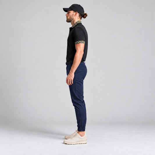A man in profile wearing a black short-sleeve polo shirt with patterned trim on the collar and sleeve, navy blue trousers, and beige sneakers, standing against a light gray studio background.