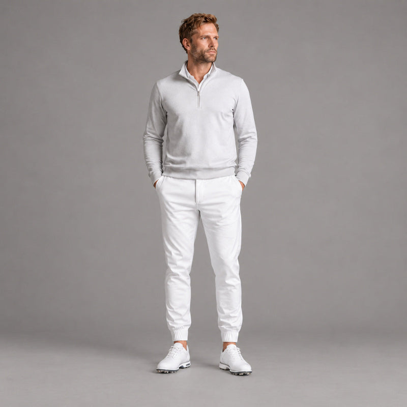 A man modeling a light gray quarter-zip pullover sweater and white jogger pants with white golf shoes, standing against a solid gray studio background.