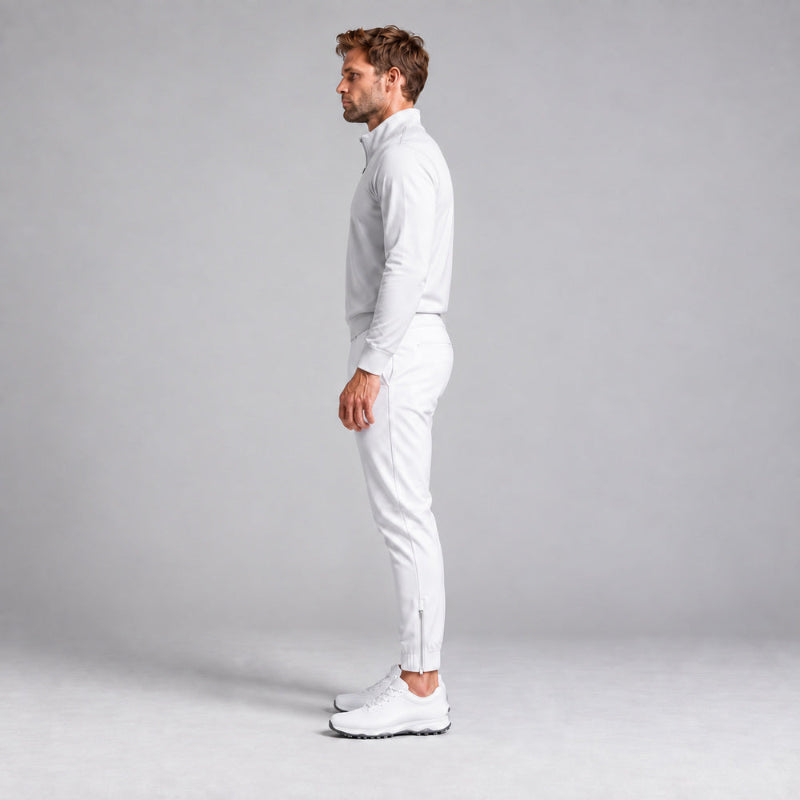 A man in a light grey long-sleeve quarter-zip pullover and white joggers stands in profile against a light gray studio background, wearing white athletic shoes.