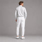 A man is shown from behind wearing a light gray long-sleeve quarter-zip pullover and matching white jogger pants with white golf shoes, set against a solid gray studio background.